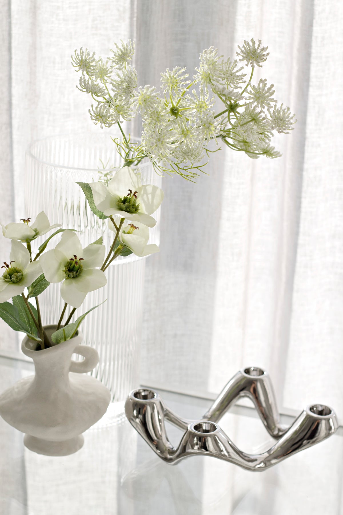 Silver candle holder on a windowsill beside a white jug of blossom and a ribbed vase