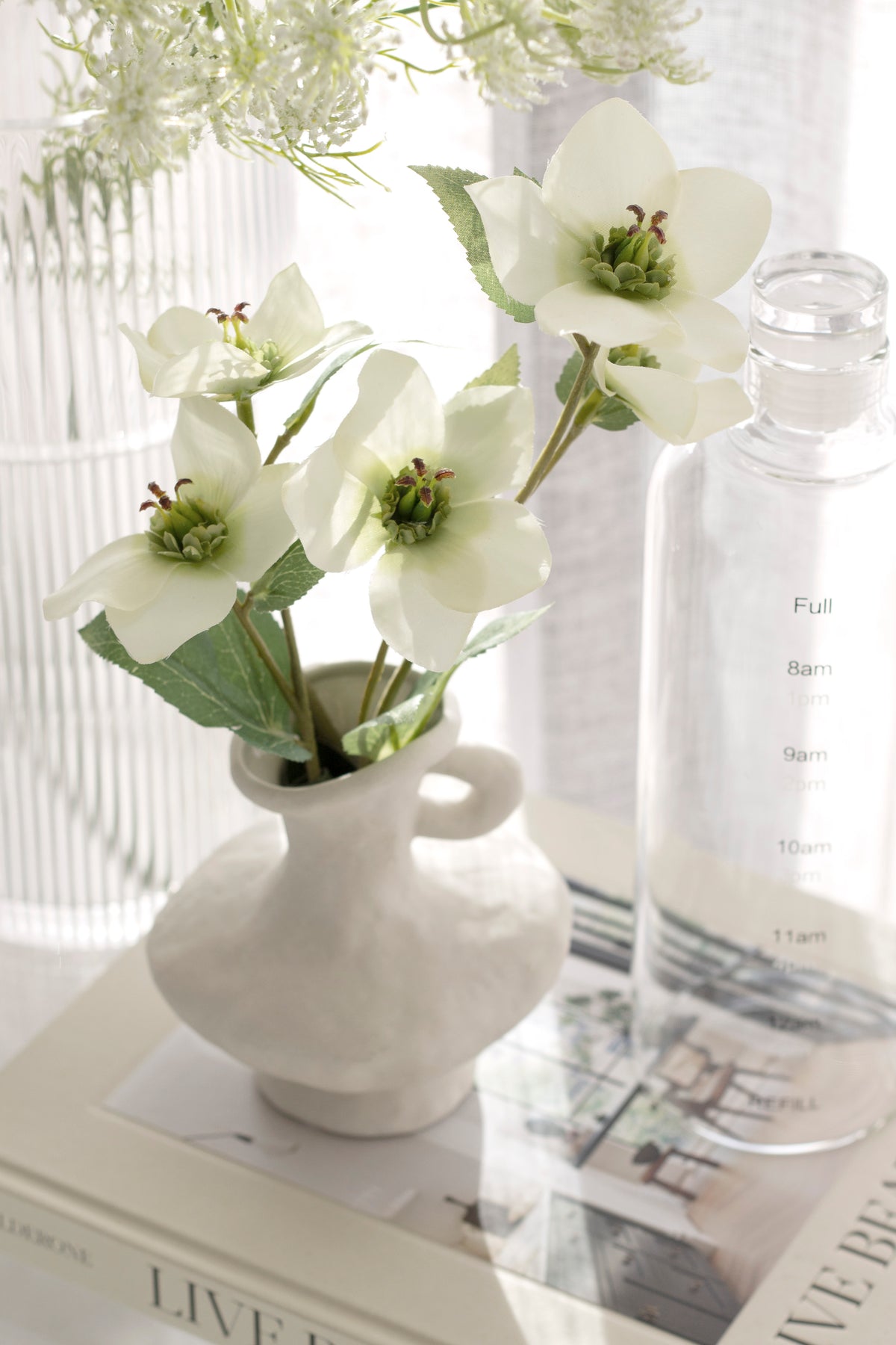 Cream hellebores in a grey vase with a handle beside a ribbed glass