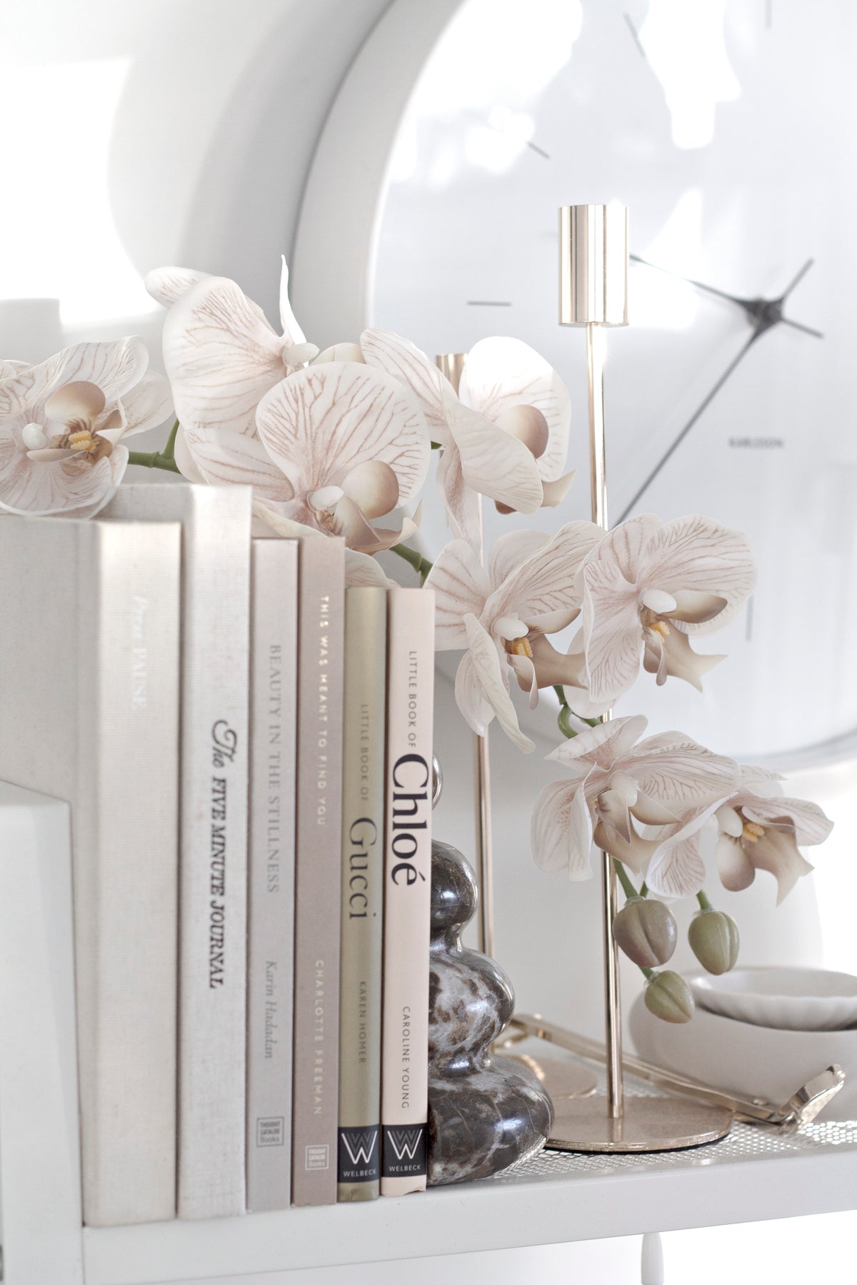 Orchids behind a row of upright books on a shelf with a brass clock and round mirror