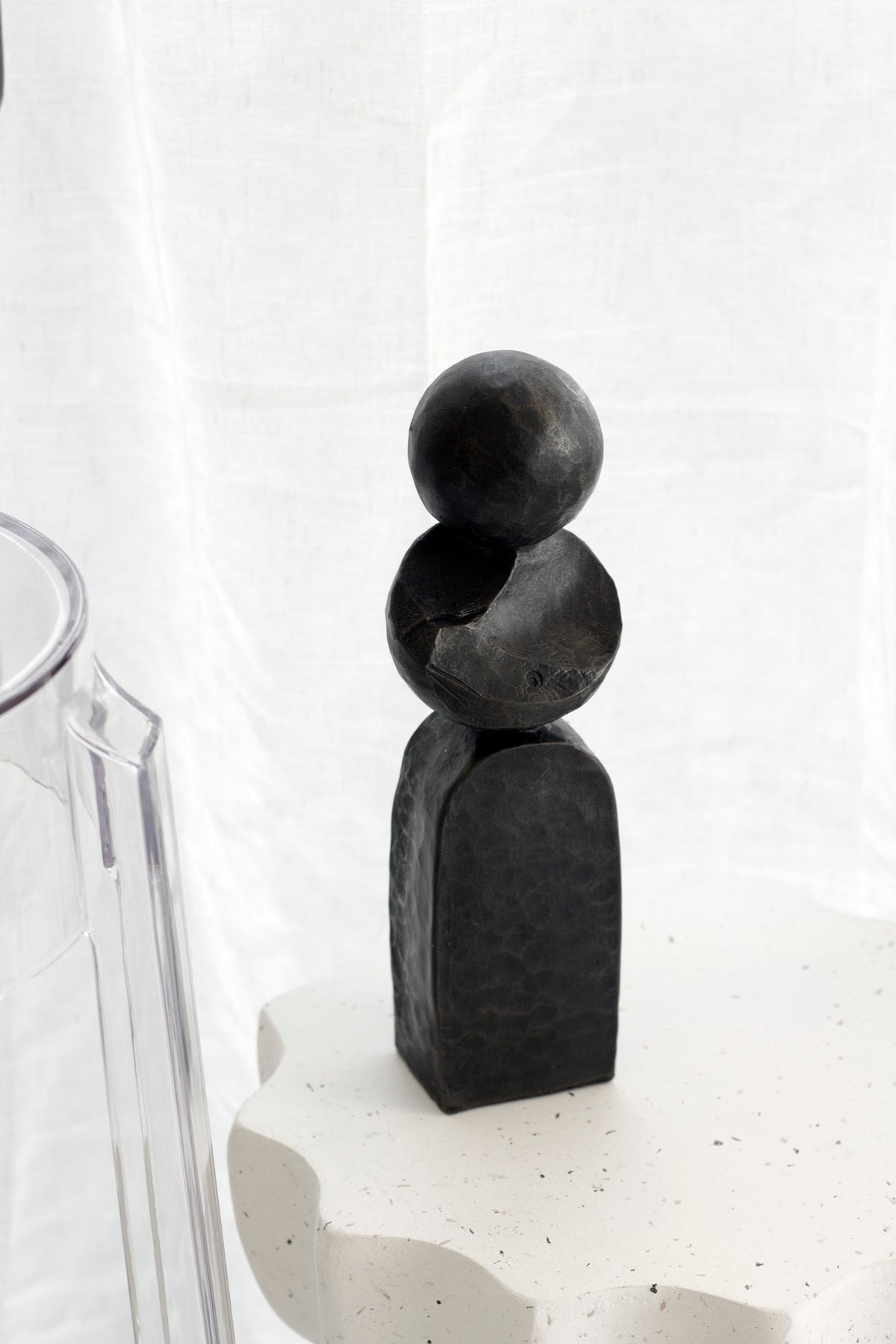 Small black Pillar sculpture on a speckled plinth beside sheer linen