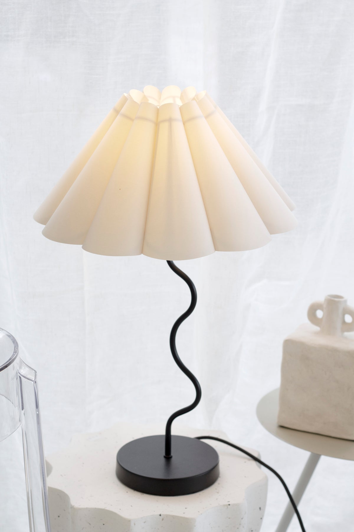 Black Wavy table lamp switched on, its pleated shade glowing warm against a grey backdrop