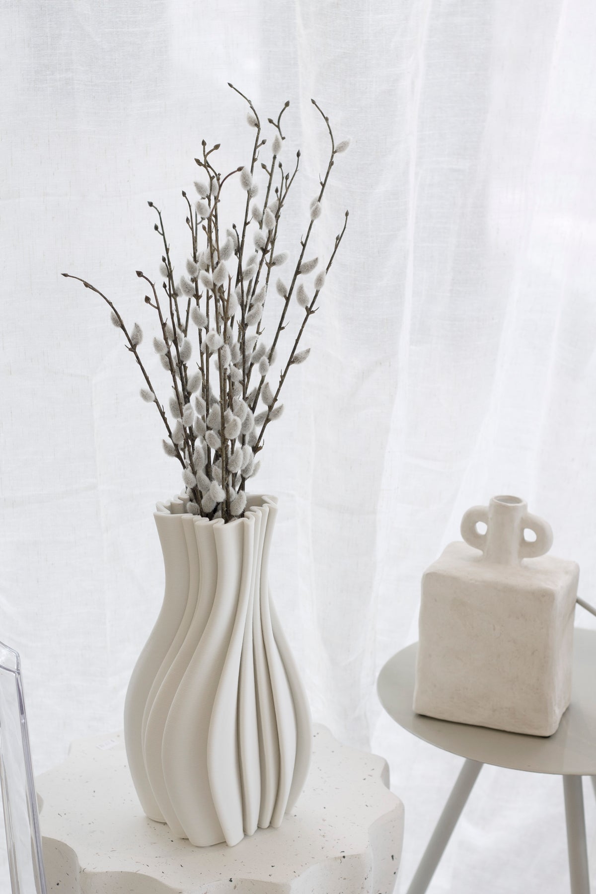 Faux pussy willow in a white wavy vase on a side table by a sheer curtain