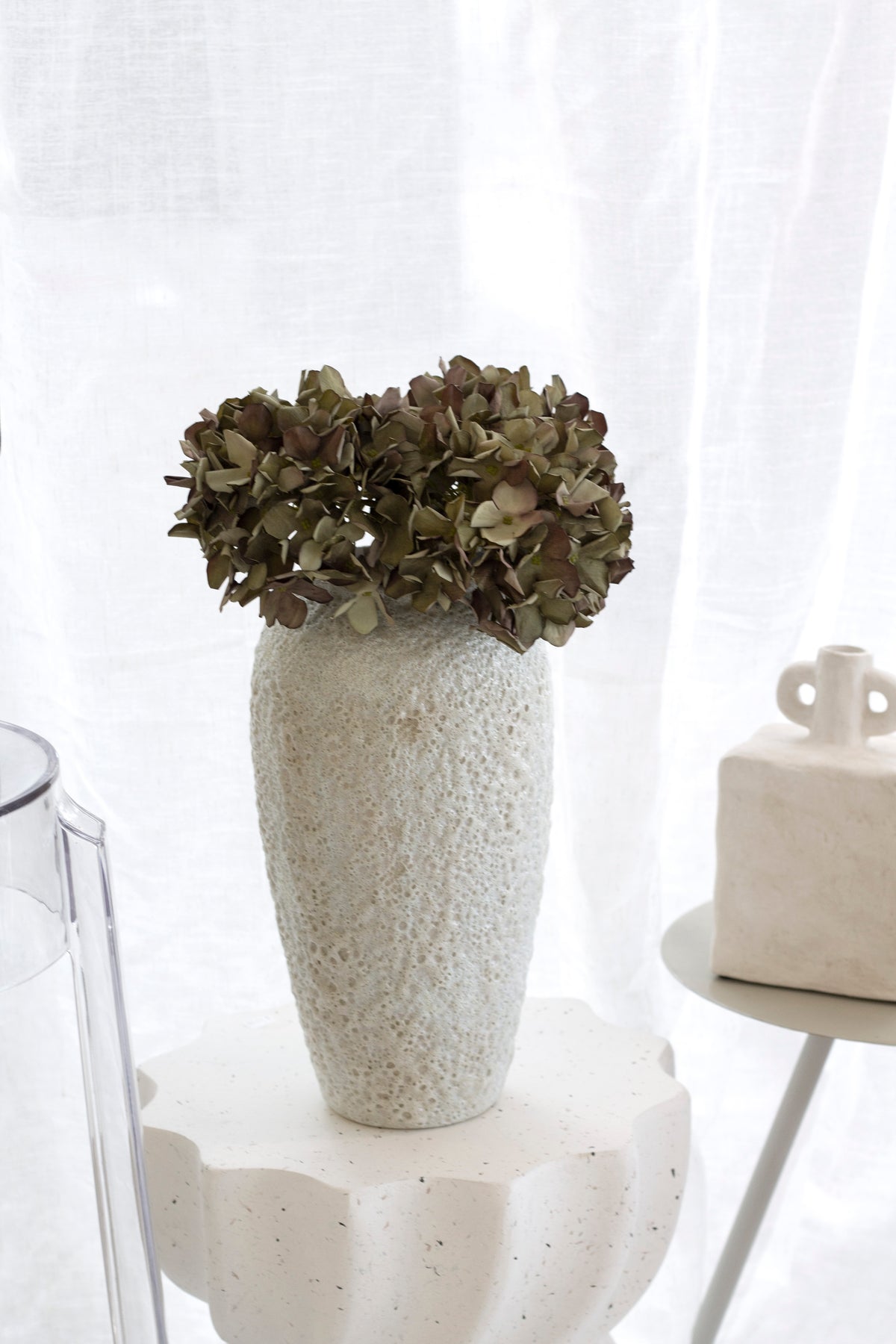 Tall Seafoam vase of dried hydrangea on a scalloped plinth, a glass cylinder vase alongside