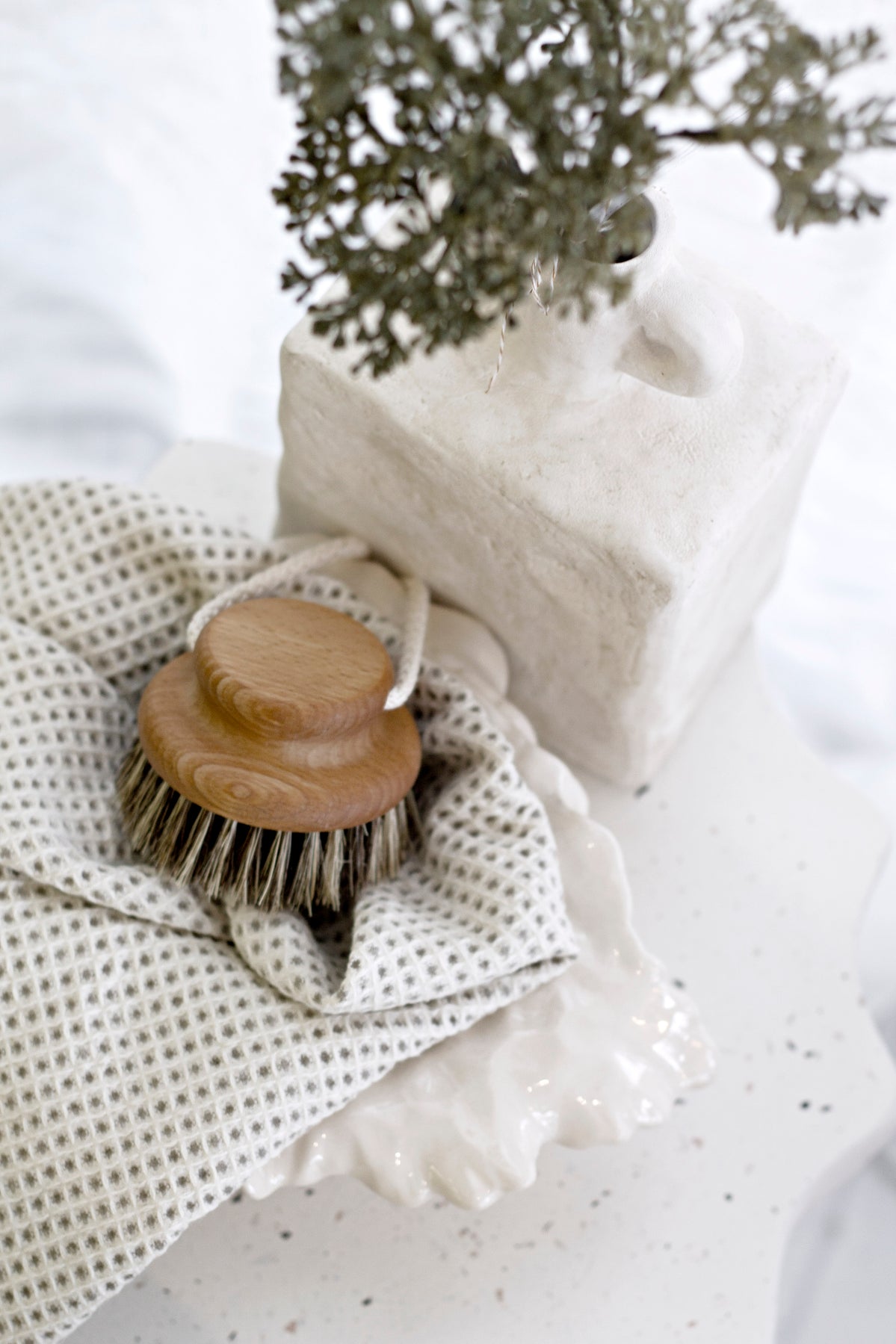 Natural body care scene featuring a wooden bristle brush resting on a textured waffle towel, beside a sculptural white ceramic vase with dried foliage, styled on a soft, speckled surface