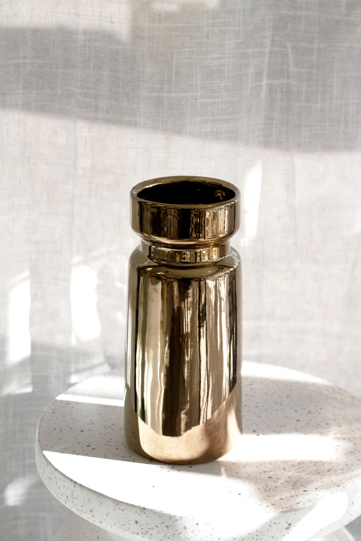 Gold cylinder vase on a white speckled terrazzo plinth in sunlight, sheer curtain behind