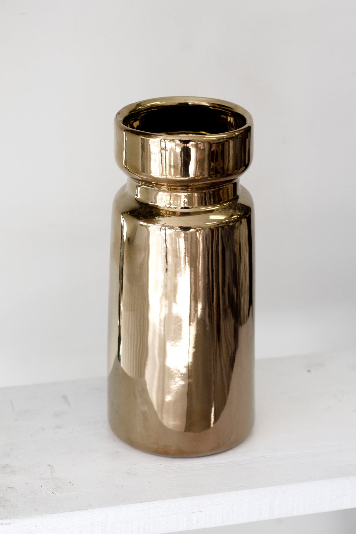 Gold cylinder vase on a white painted ledge, its mirrored surface reflecting the pale wall