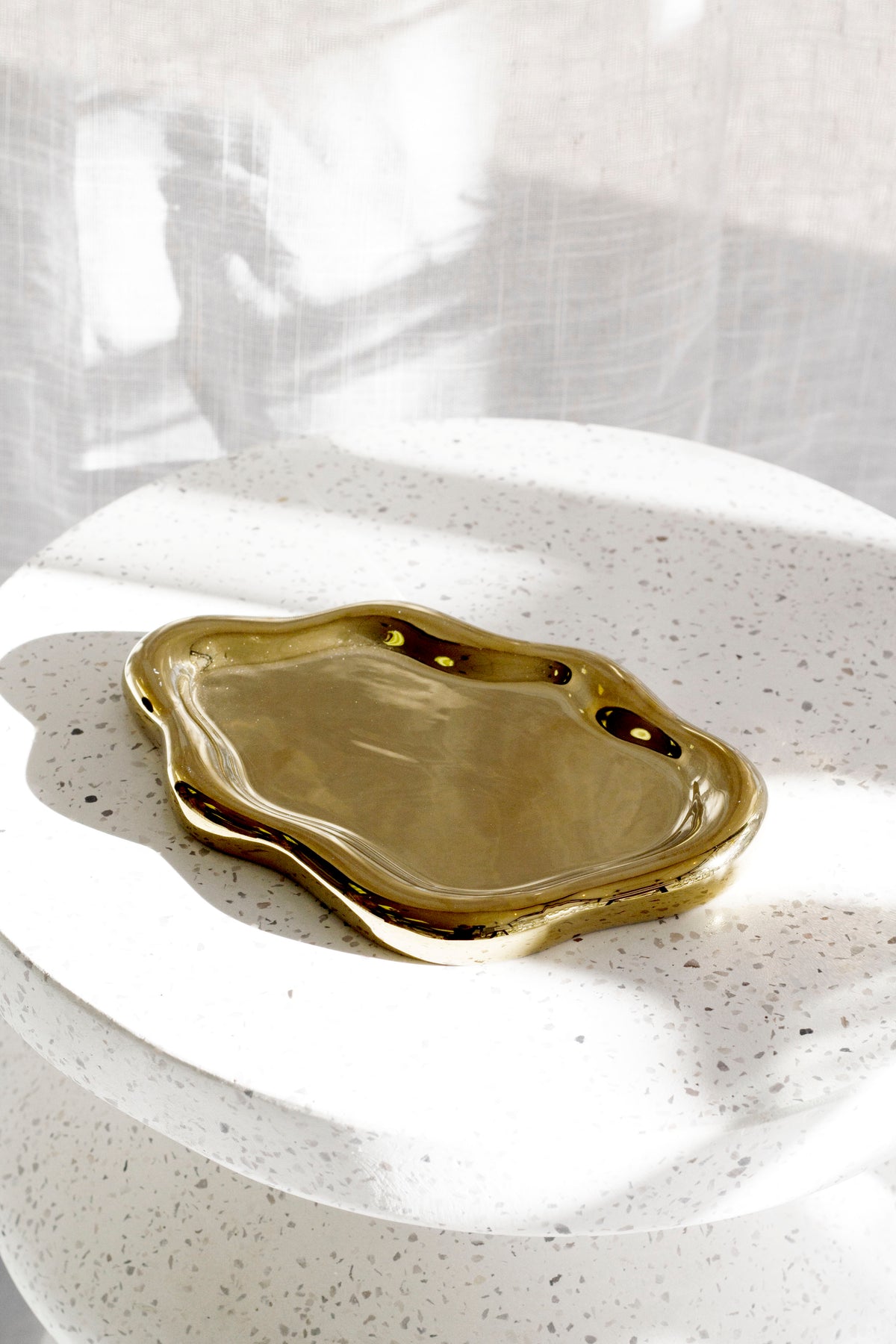 Gold cloud tray on a white speckled terrazzo plinth with a sheer curtain behind