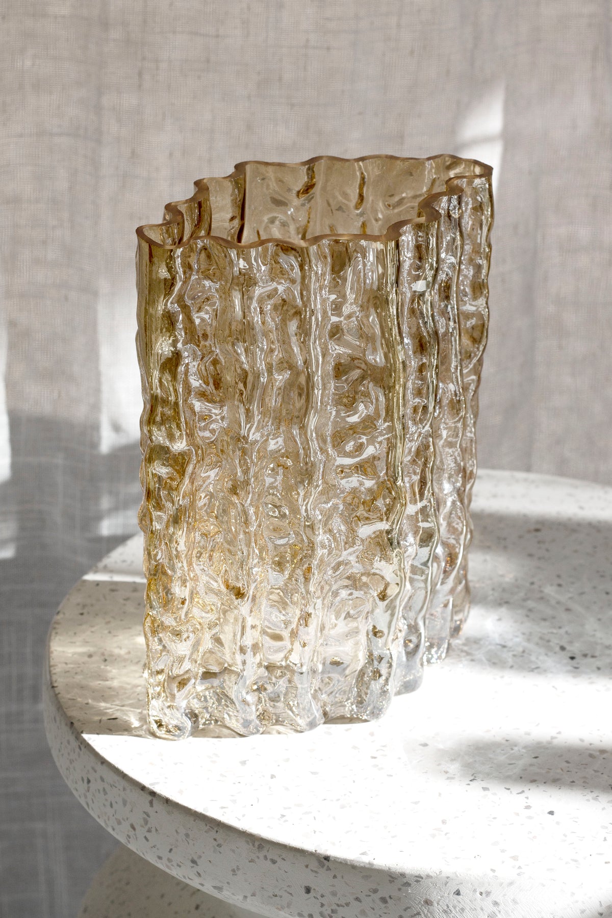 Gold Glacier vase on a stone pedestal in warm sunlight against a linen curtain