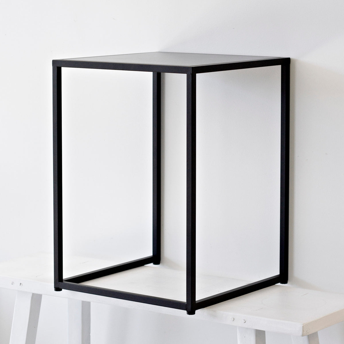 Sara black side table on a pale bench against a white wall, open frame showing through