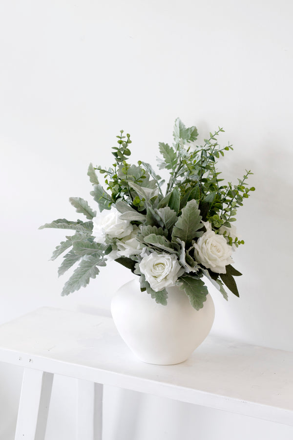 Artificial silver dollar eucalyptus spray styled in a white matte vase with soft white roses and textured green foliage on a white bench against a minimalist wall.