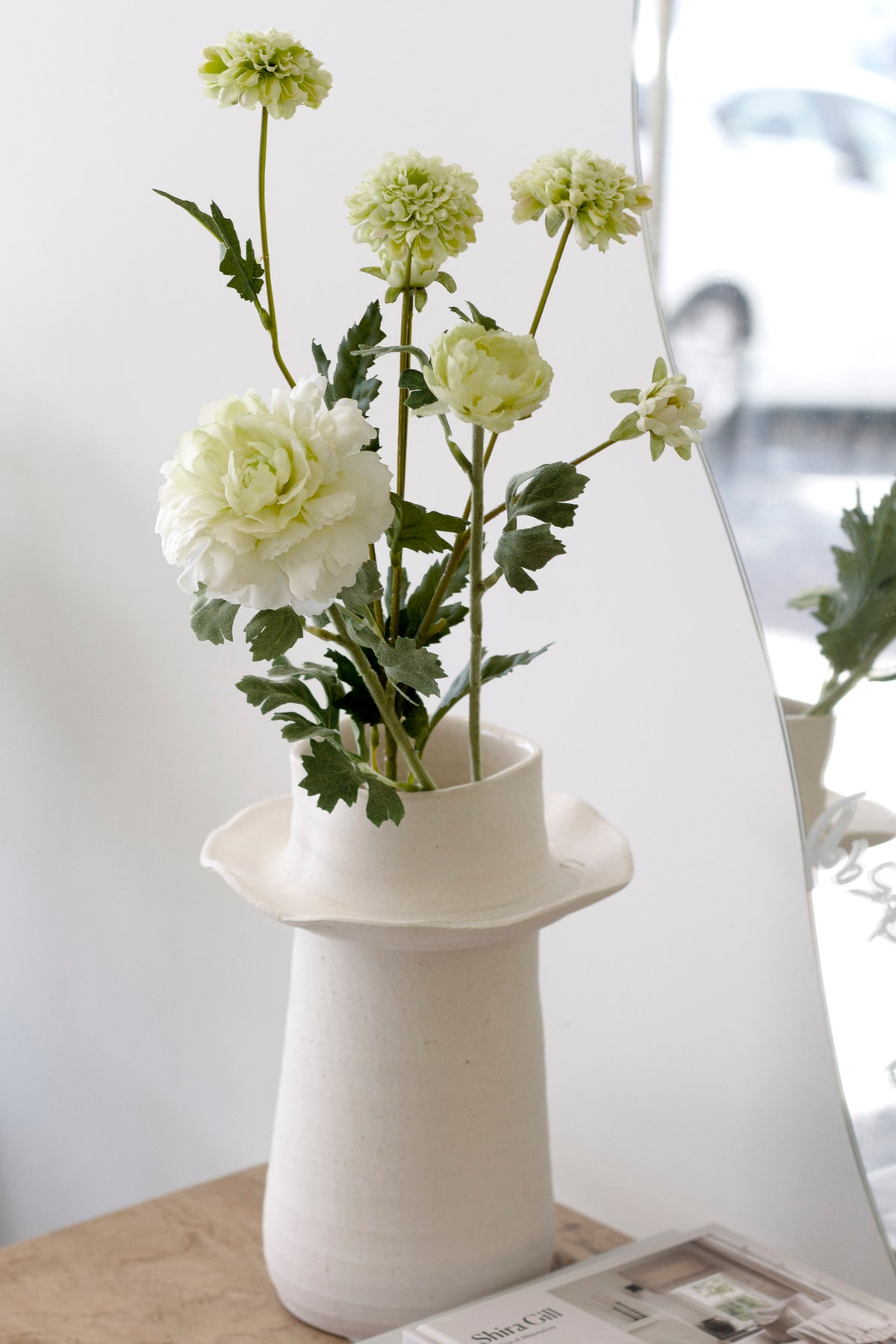 White and green pompom mum stems arranged in a cream sculptural vase on a timber surface