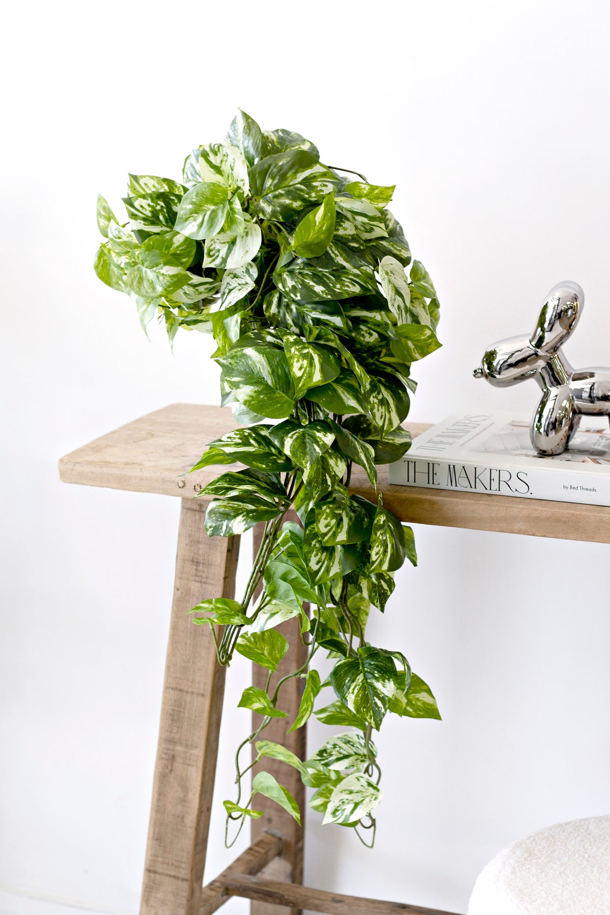 Pothos trailing from a timber shelf above a boucle stool, books and a silver ornament alongside
