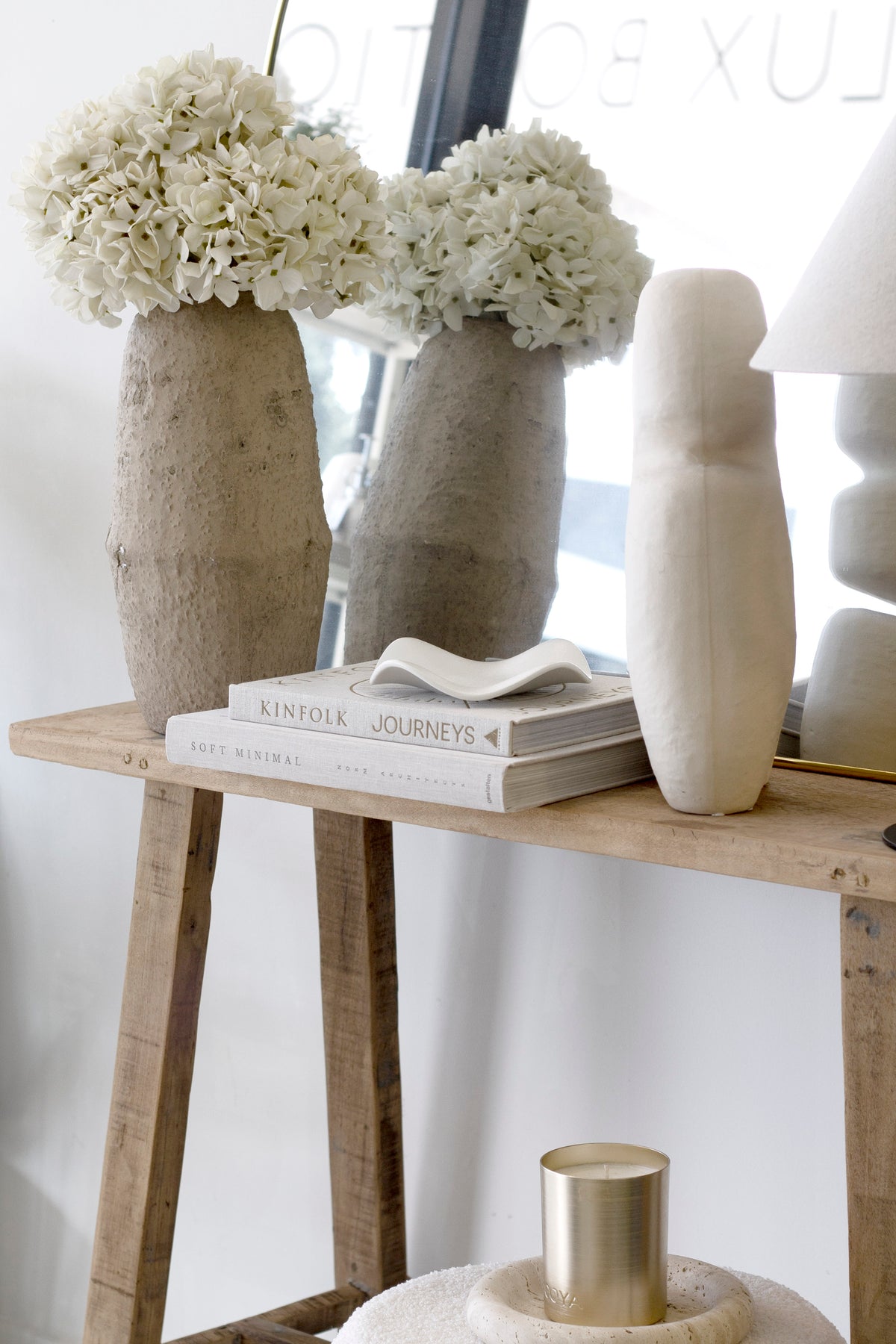 Two textured vases of white hydrangeas on a timber console with books and a pale lamp