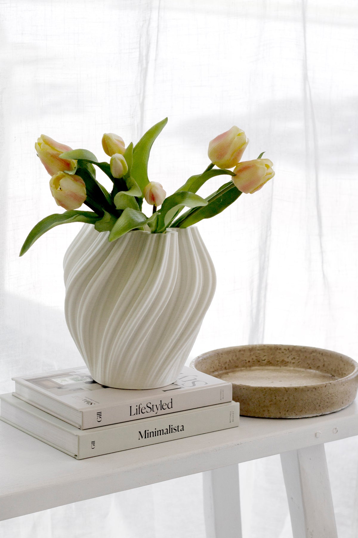 Peach faux tulips in a swirled cream vase on stacked books beside a woven bowl