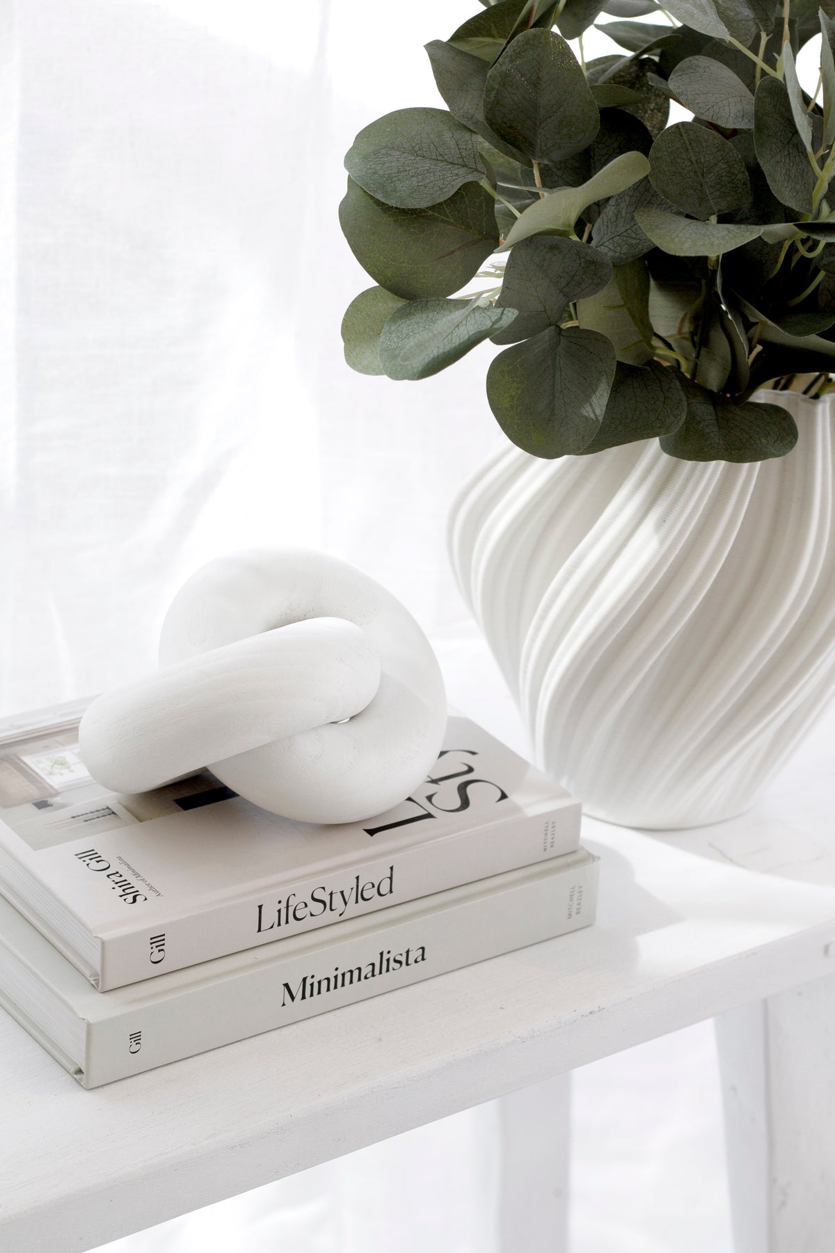 White Amelie fluted vase with eucalyptus beside a white knot ornament on stacked books
