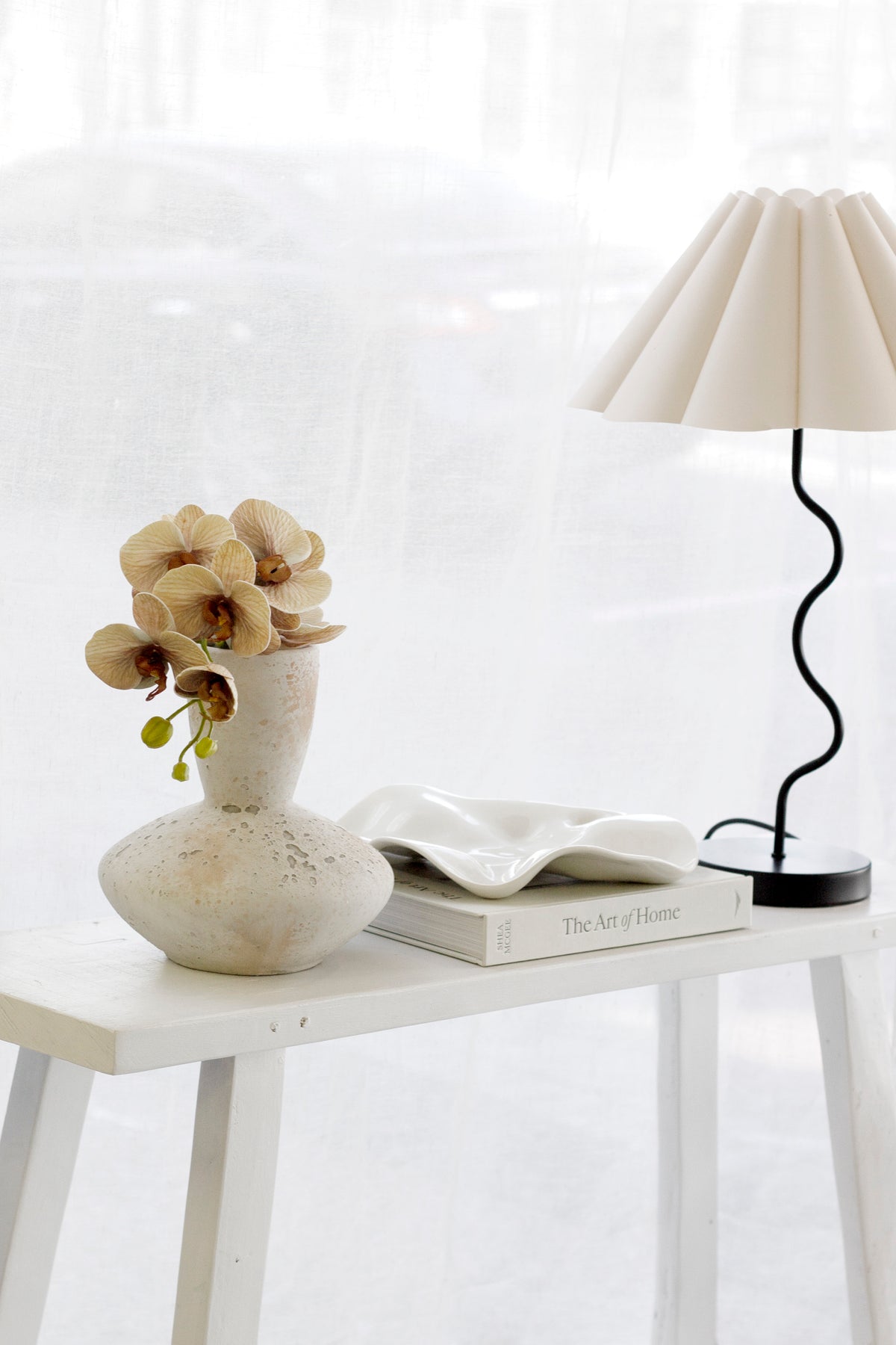 The Art of Home on a white table under a sculptural tray, a stone vase of orchids and a wavy lamp