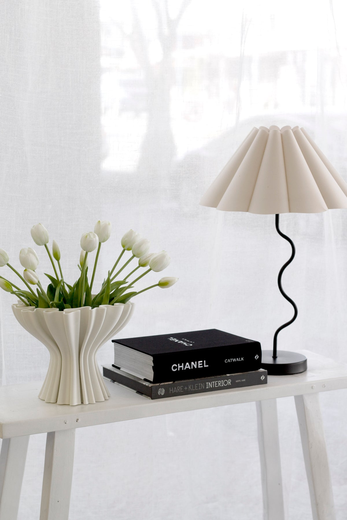 Sculptural white vase of white faux tulips on a bench beside coffee table books and a pleated lamp