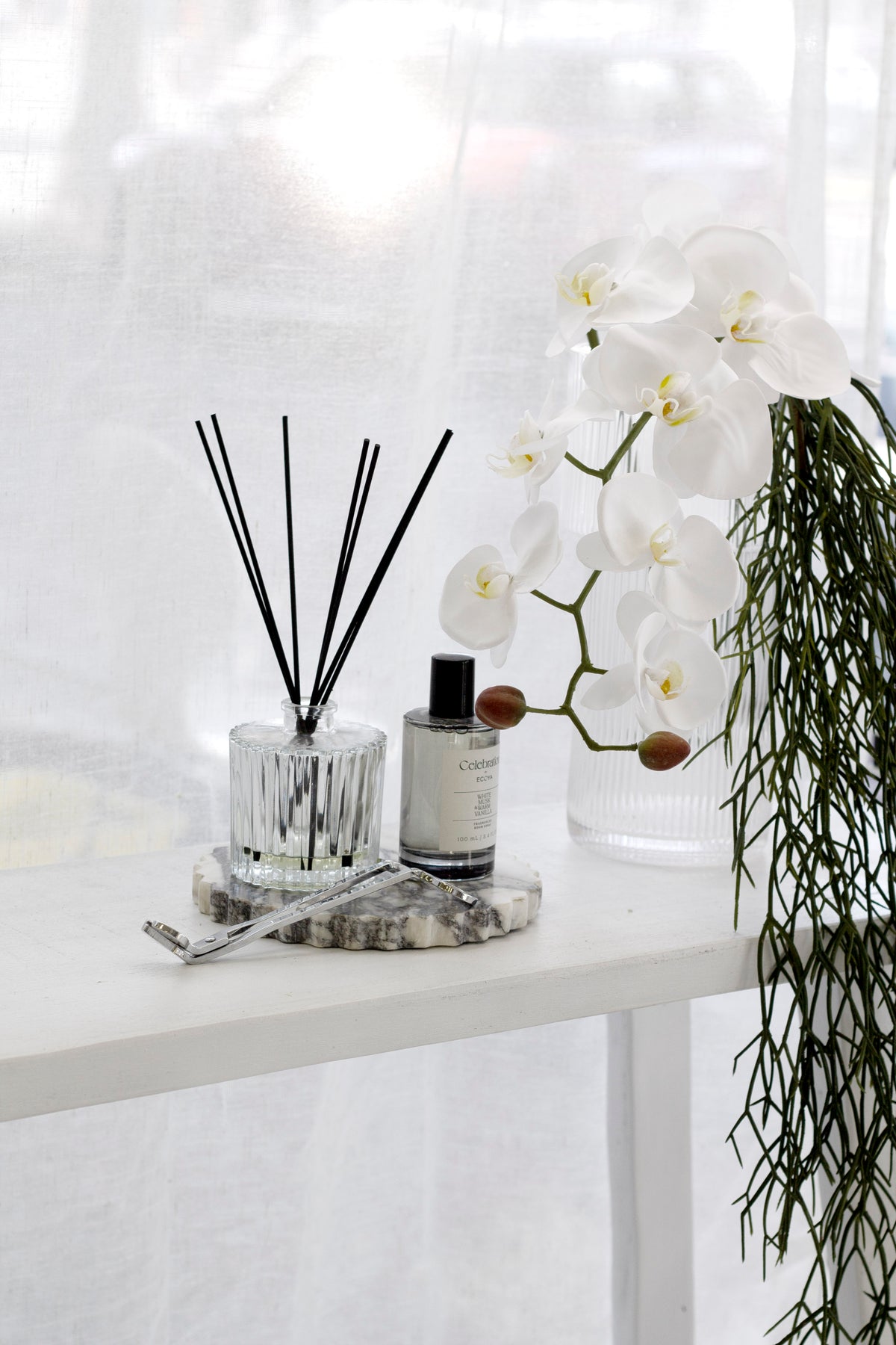 Celebration diffuser on a white console with a white orchid and a bottle of hand wash