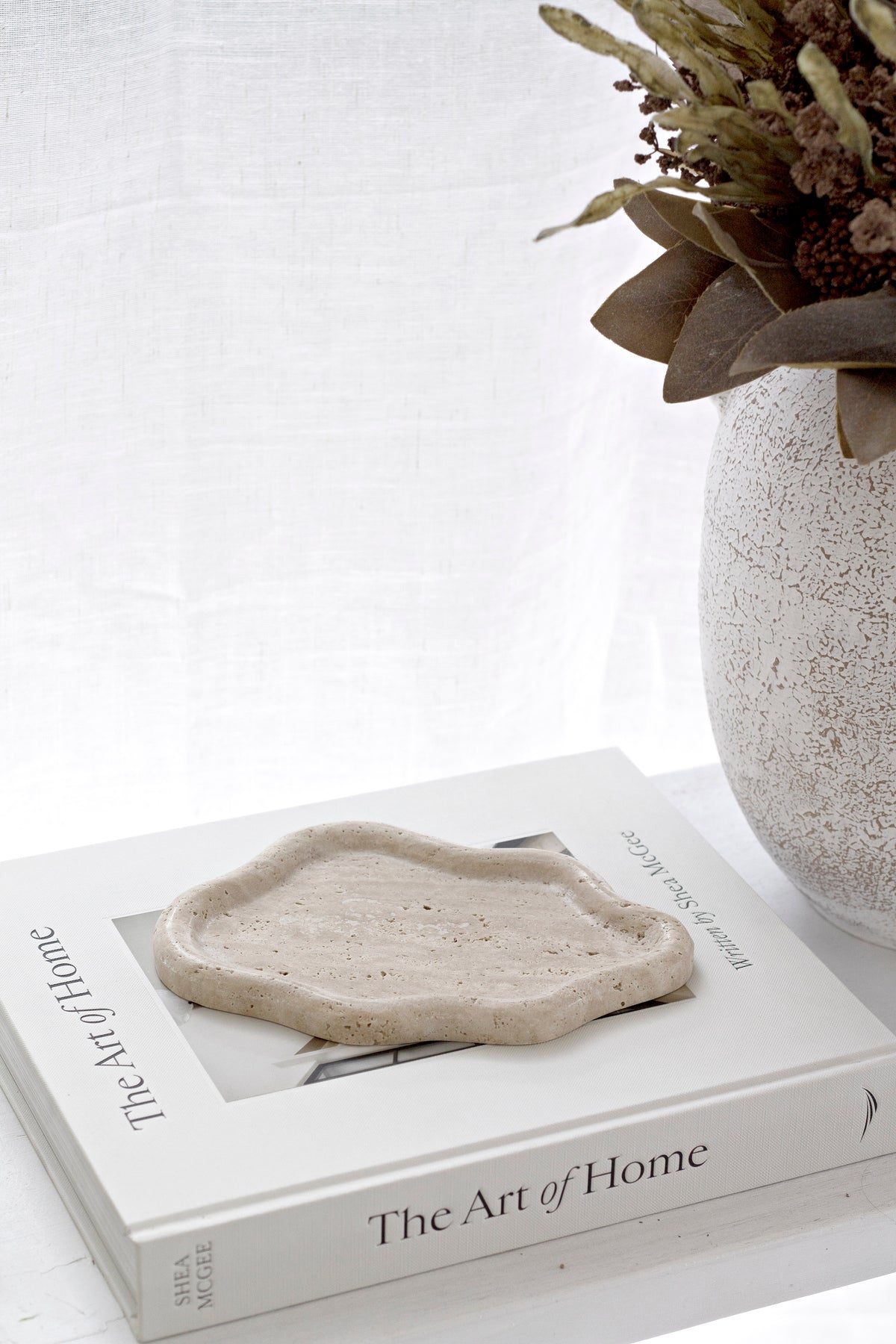 Travertine catchall tray on a stack of books beside a speckled vase of dried stems