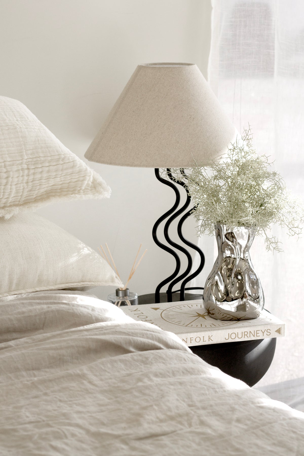 Surge table lamp on a bedside beside soft bedding and a vase of flowers
