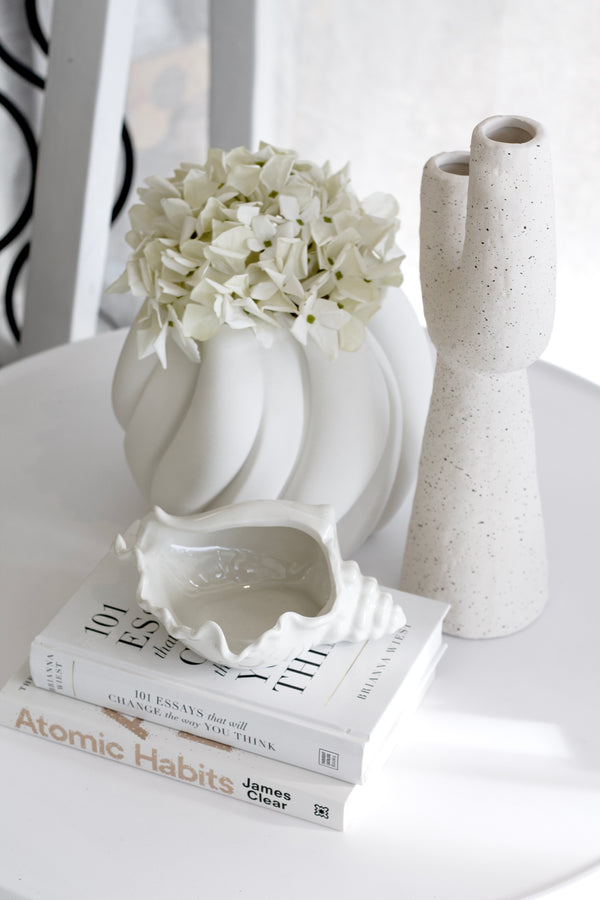 Neutral-toned styled vignette with Atomic Habits by James Clear stacked under 101 Essays That Will Change the Way You Think, topped with a white ceramic shell dish, sculptural vases, and white faux hydrangeas.

