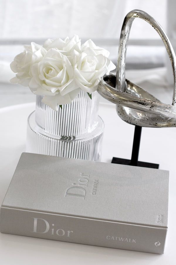 Silver metallic loop sculpture on a black stand styled beside fluted glass vase with white roses and a Dior Catwalk book on a white tabletop.