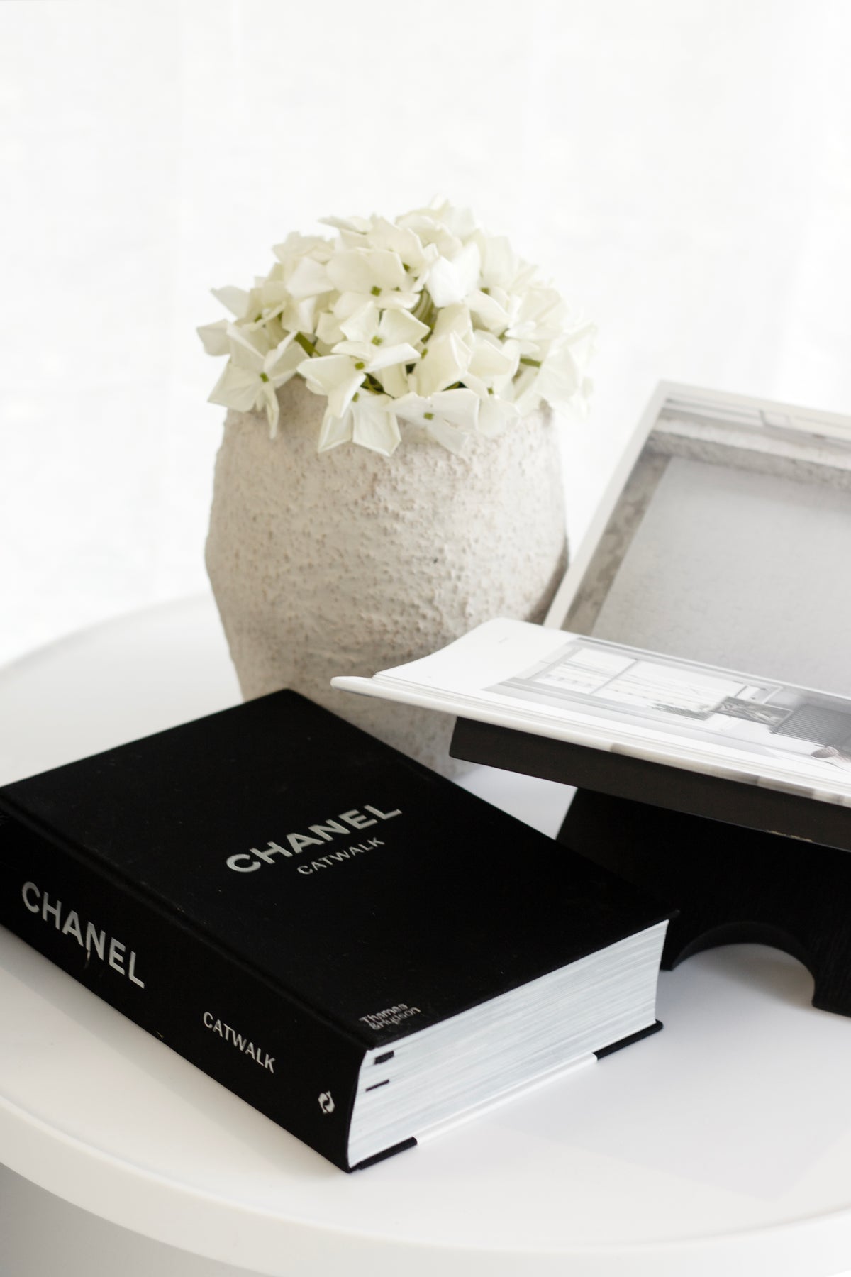 Black wooden Como Book Stand styled on a round white table holding an open book, next to a textured vase with white faux hydrangeas and a black Chanel Catwalk book.