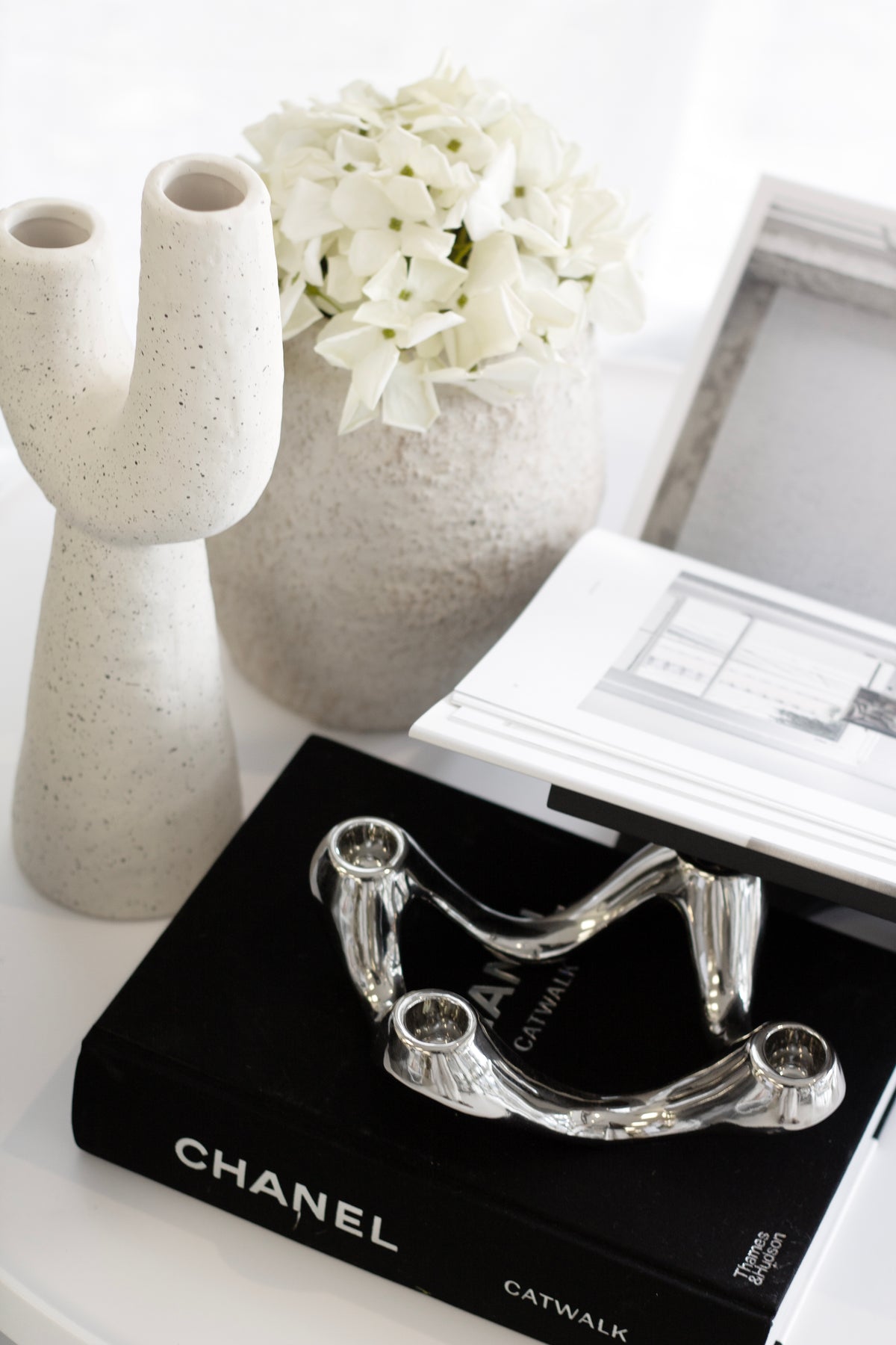 Silver candle holder on a black fashion book beside two textured white vases and hydrangea