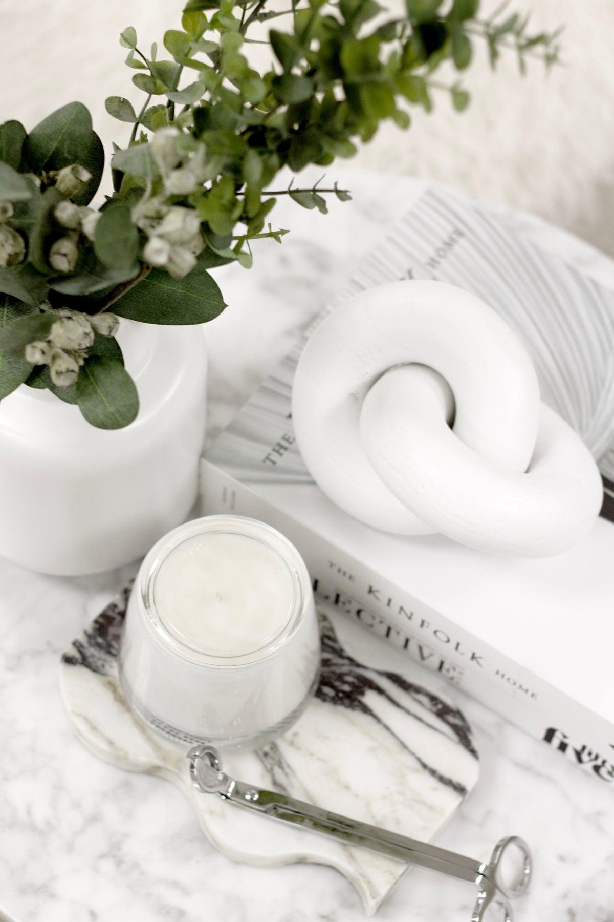 White wooden knot sculpture with a lit candle, eucalyptus and scissors on white linen