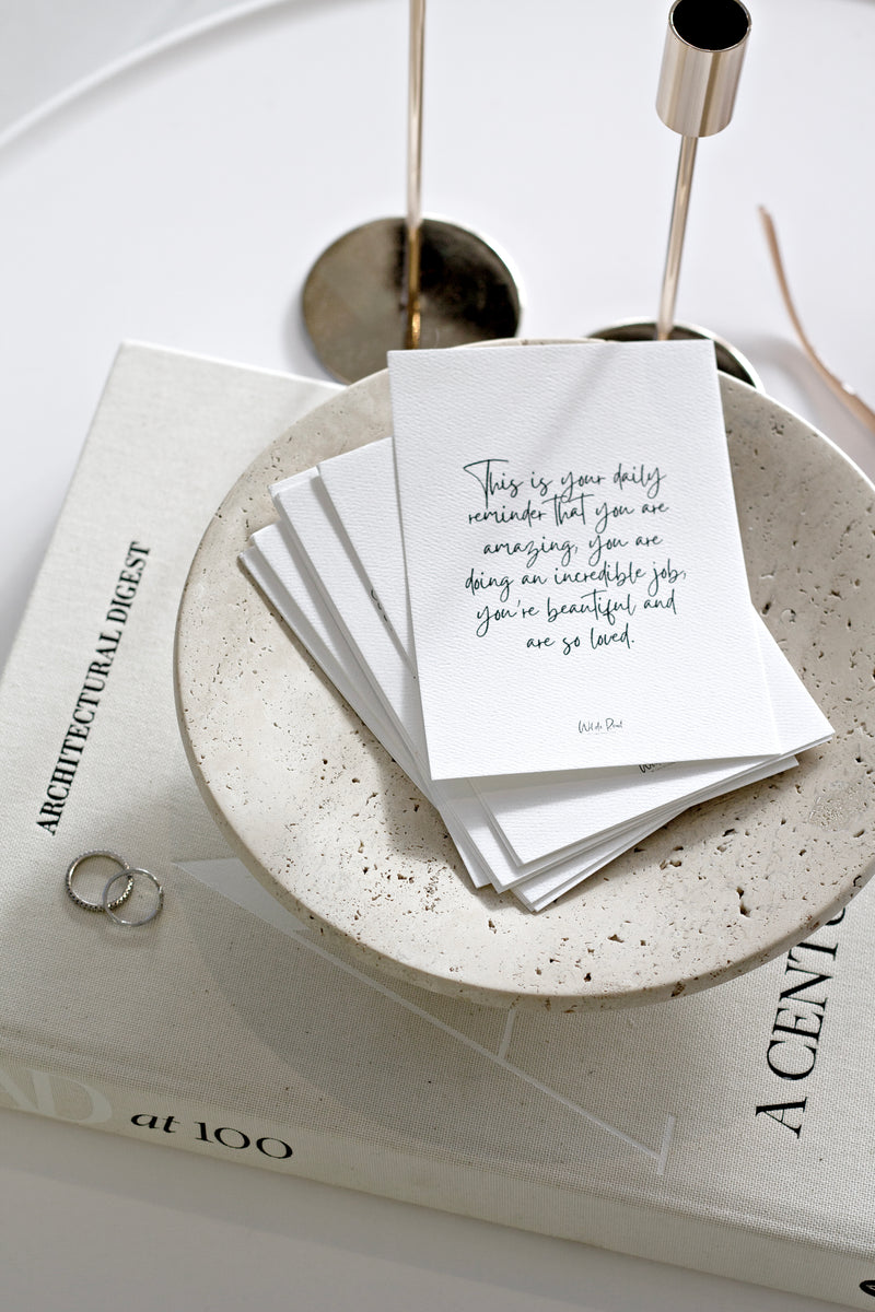 A textured stack of daily affirmation cards in a terrazzo-style bowl, with the top card reading: “This is your daily reminder that you are amazing, doing an incredible job, you’re beautiful and so loved.”
