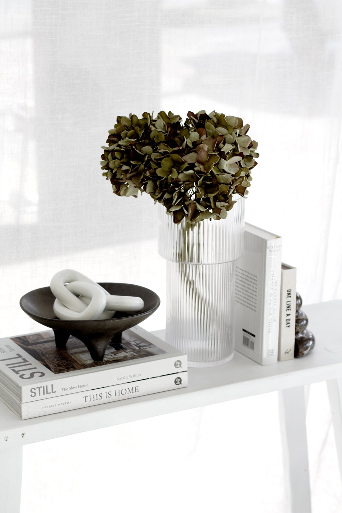 Ribbed glass vase of dried hydrangea on books beside a dark dish holding a white knot