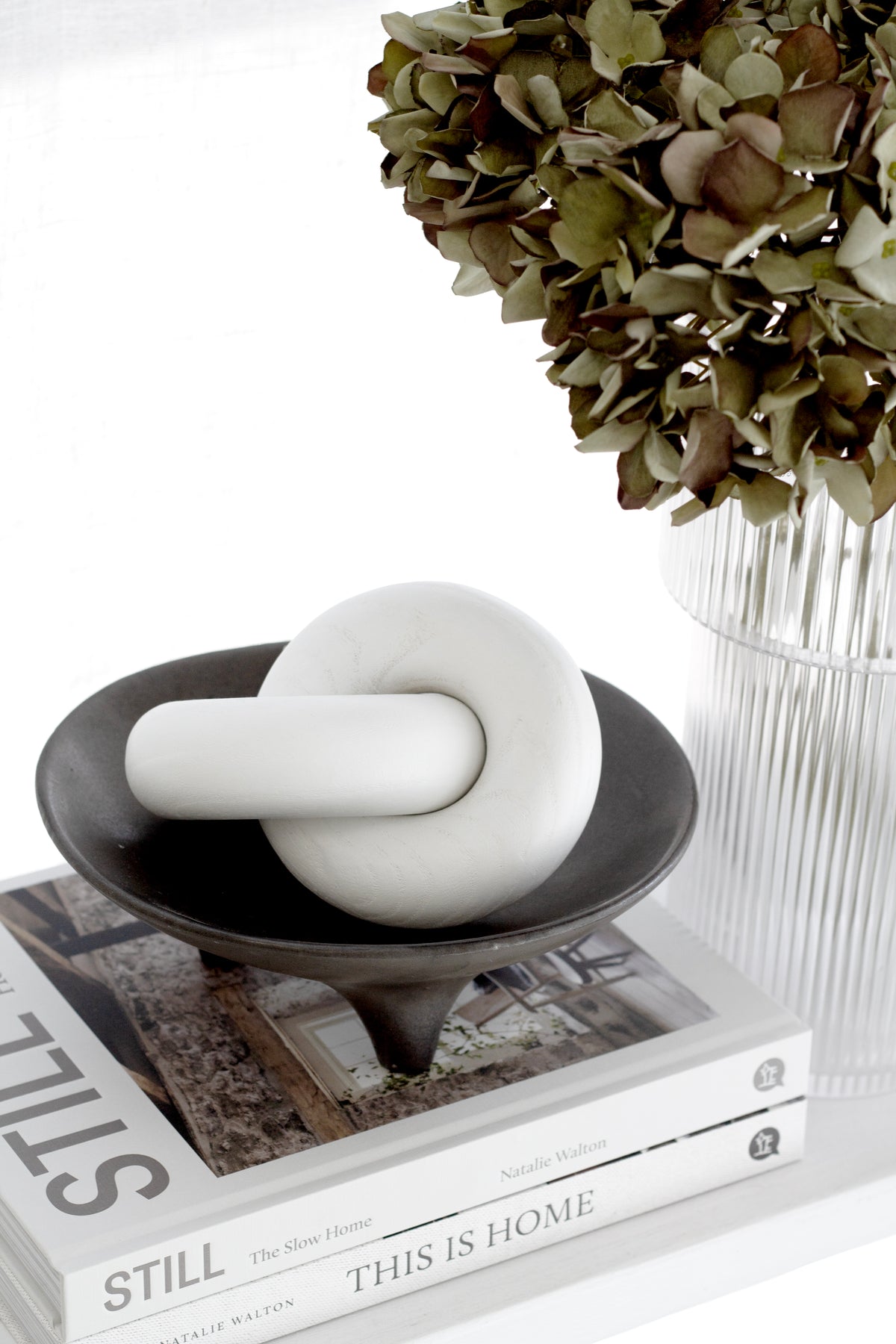 White wooden knot sculpture in a dark tripod dish on books, dried hydrangea behind