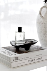 Matte black ceramic dish with sculptural feet holding a perfume bottle and ring, styled on stacked coffee table books beside a rustic white vase.