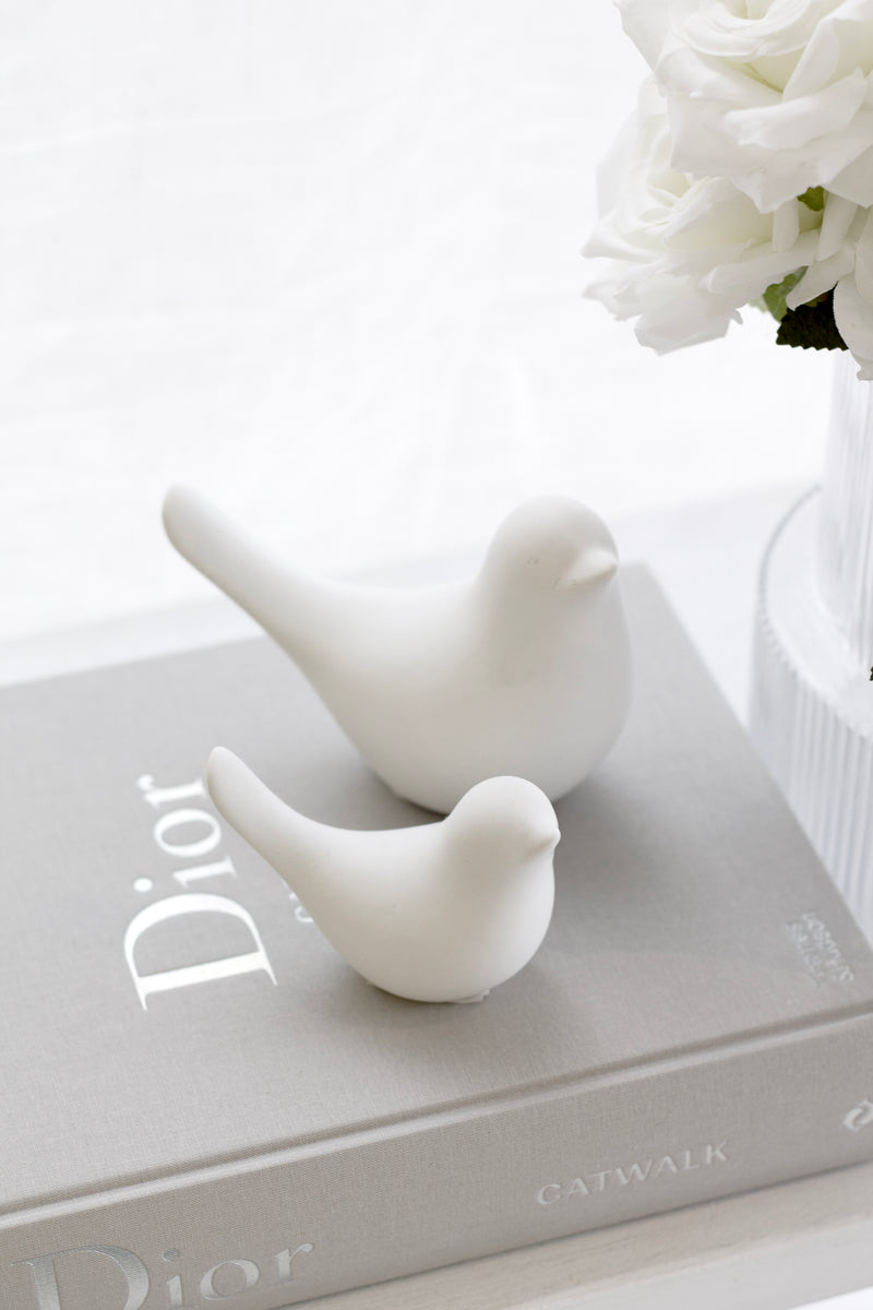 Two matte white ceramic dove sculptures styled atop a grey Dior book, beside a vase of white roses for a peaceful and minimal look.