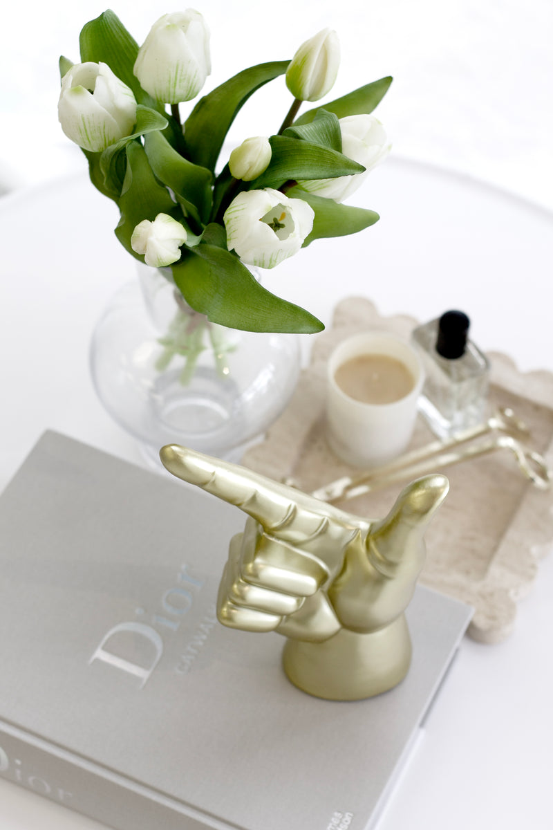 Glossy brass resin hand sculpture styled on a Dior Catwalk book beside a vase of white tulips and a minimalist candle.