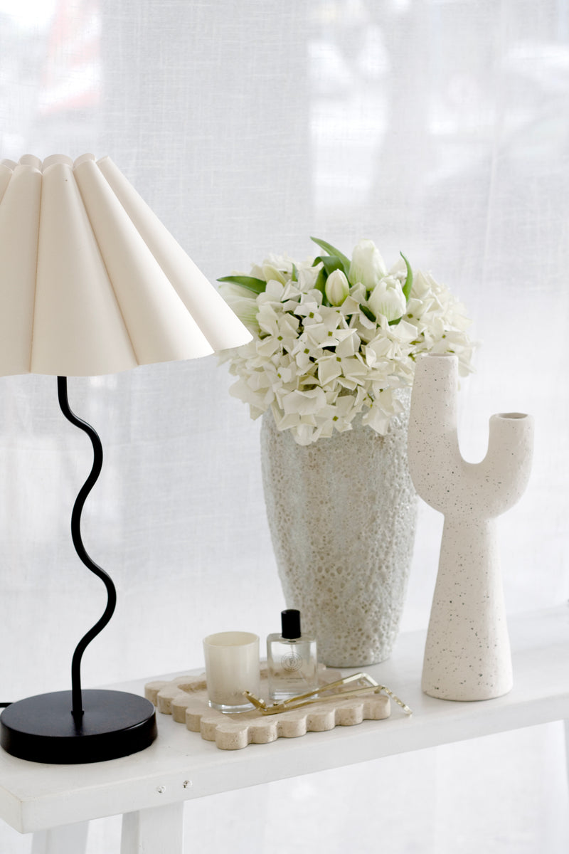 White artificial Grandiflora Hydrangea stems arranged in a textured vase on a white console table, styled with a wavy black lamp, tray, and neutral decor.