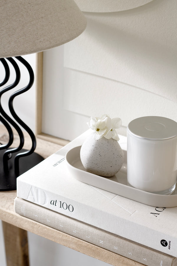 A small, rounded ceramic pebble vase with white blooms styled on a stack of books next to a candle and sculptural black lamp, creating a soft, organic vignette.