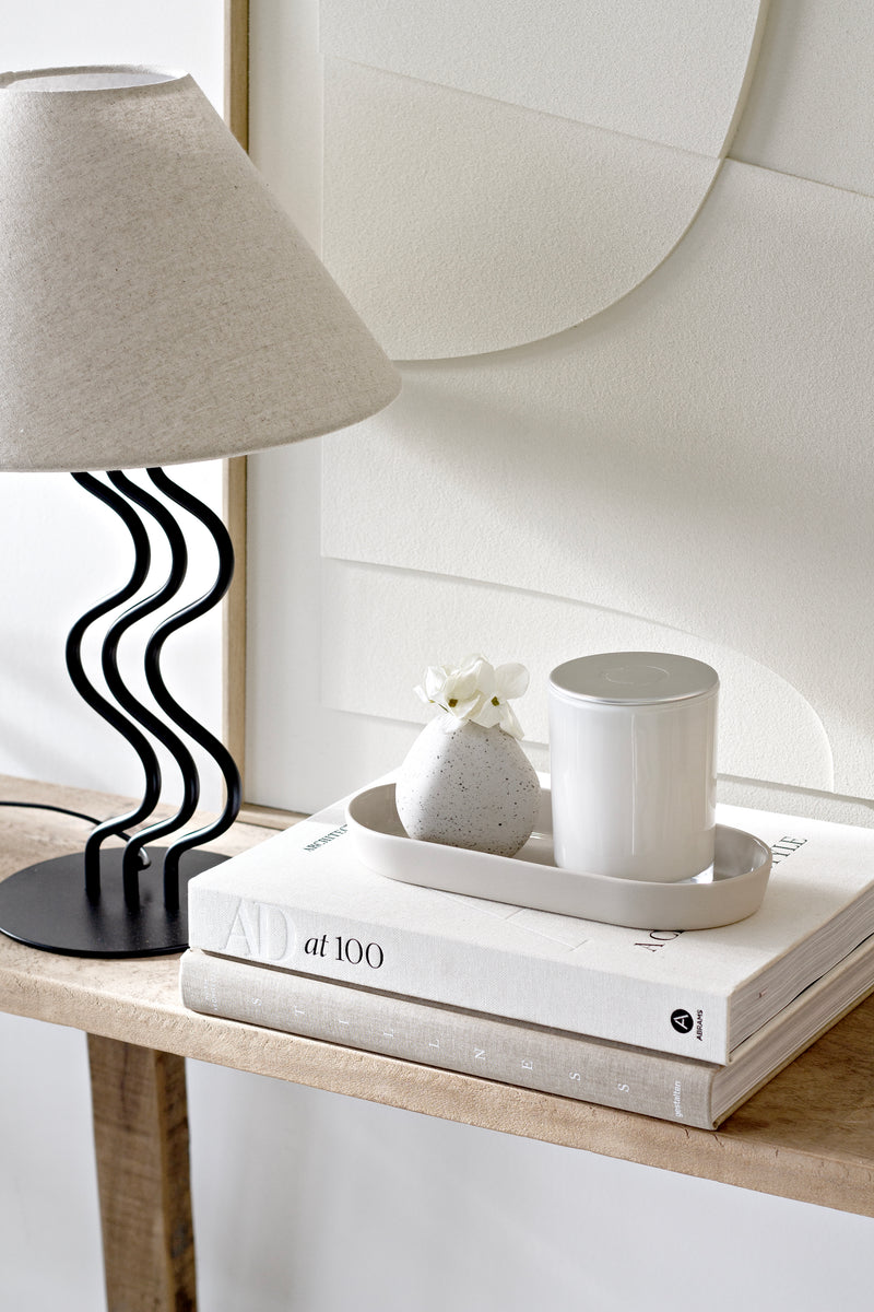 Rounded neutral ceramic vase styled with a white candle on a tray, next to books and a wavy black lamp.