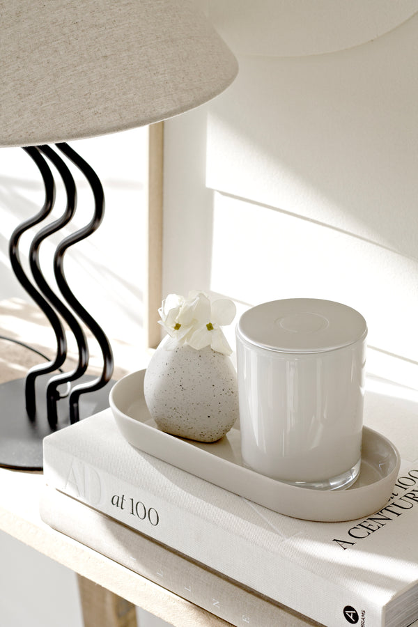 A small oval ceramic dish in soft neutral tones holding a speckled bud vase with white flowers and a minimalist white candle, styled on stacked design books next to a sculptural black lamp.
