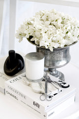 Antique-style pewter urn filled with white hydrangeas, styled on a coffee table with fashion books, a white candle, and marble accessories.