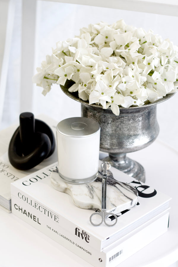 Antique-style pewter urn filled with white hydrangeas, styled on a coffee table with fashion books, a white candle, and marble accessories.