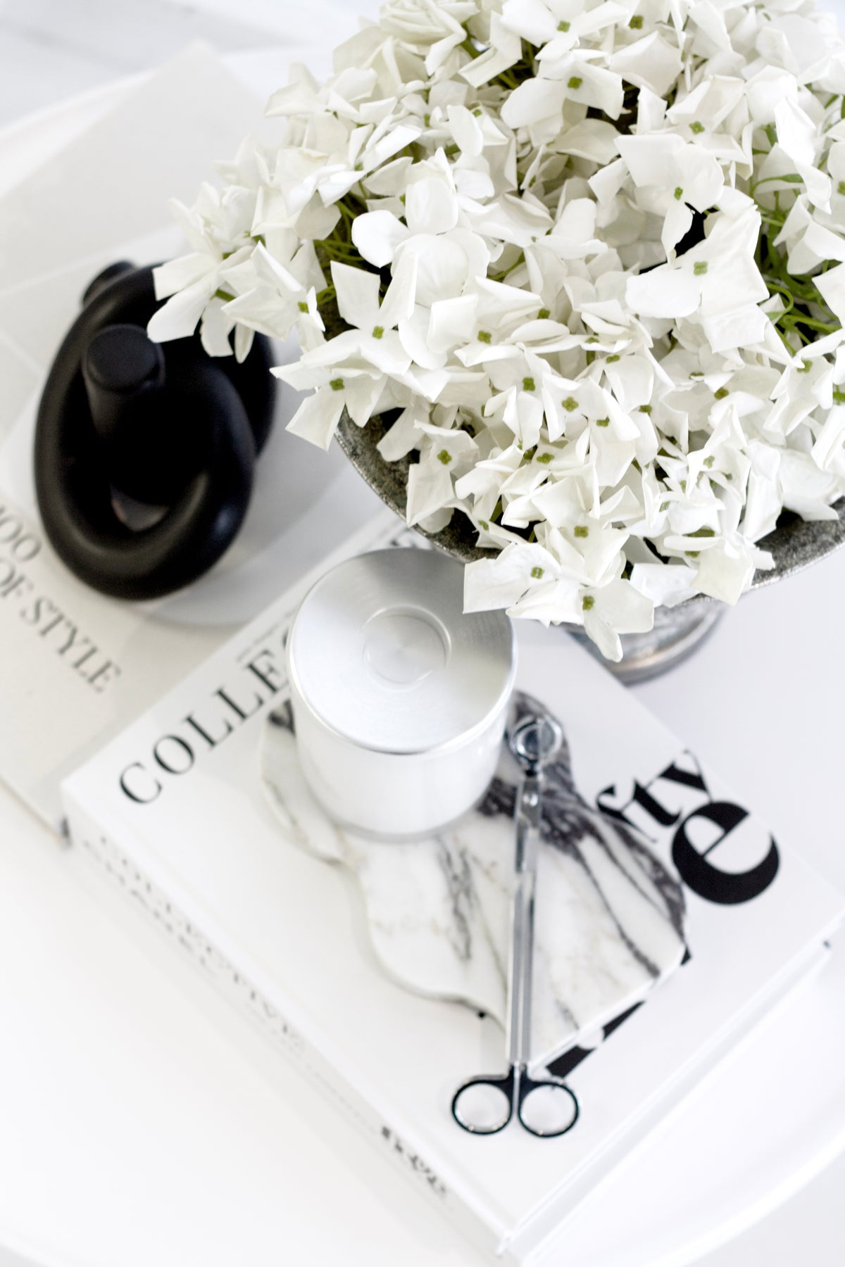 White flowers in a vase on a magazine with scissors and a bottle cap.