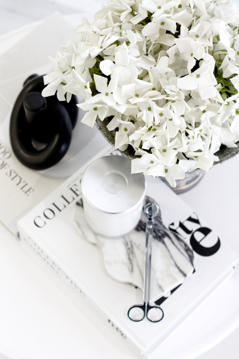 White flowers in a vase on a magazine with scissors and a bottle cap.