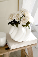 Matte white Emmy vase with sculptural curves displayed atop a Chloe book and stool, filled with soft white faux roses in a sunlit interior.