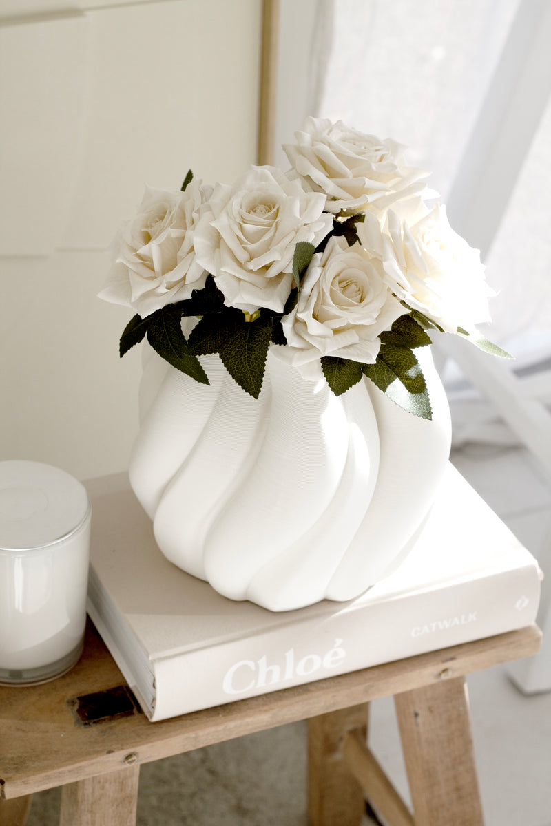 Matte white Emmy vase with sculptural curves displayed atop a Chloe book and stool, filled with soft white faux roses in a sunlit interior.