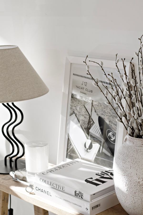 A set of soft faux pussy willow stems in a textured white vase, styled on a wooden console beside coffee table books, a sculptural black lamp, and a monochrome Chanel photo print.