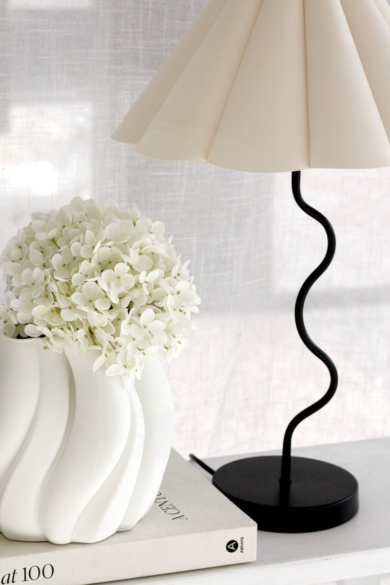 A sculptural white vase styled with a full cream artificial hydrangea stem beside a pleated cream lampshade and black curved lamp stand on a white console with books.