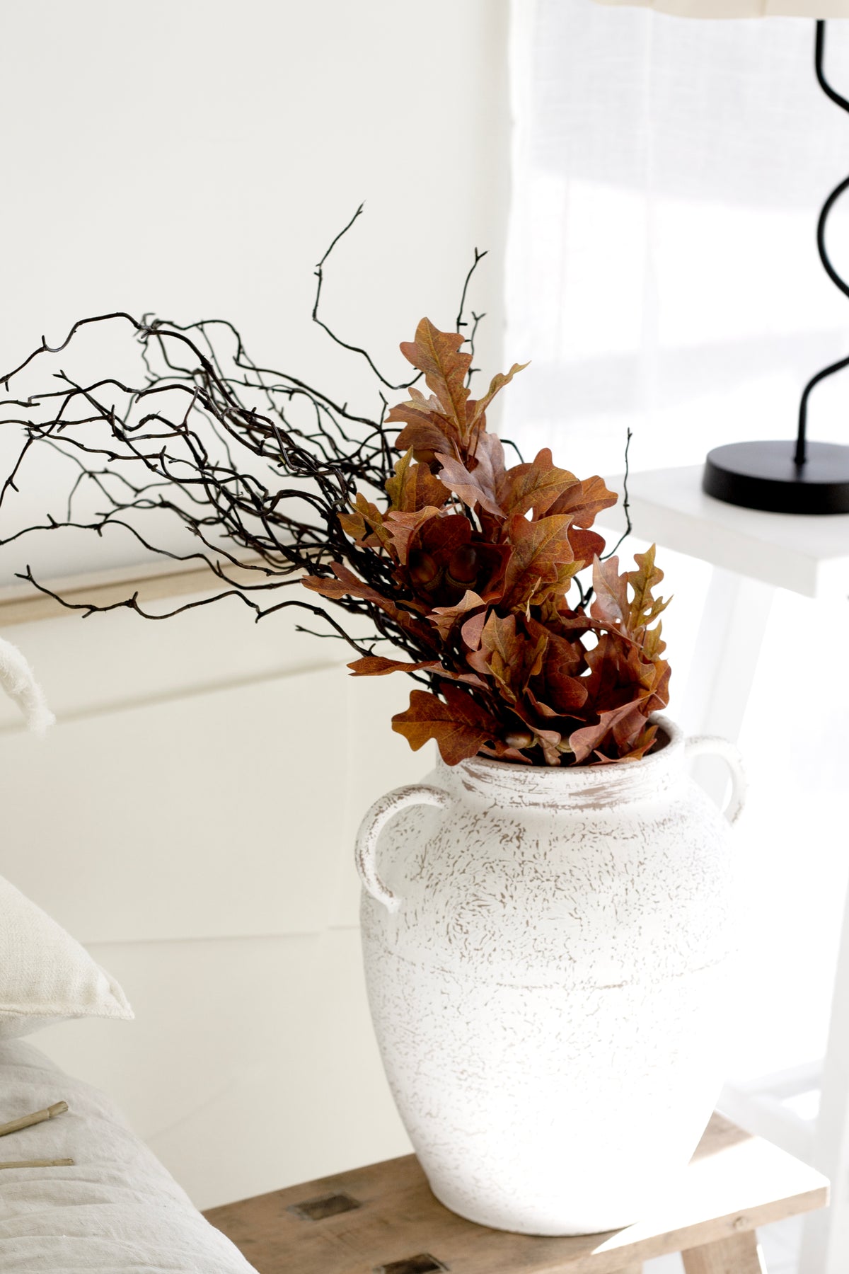 Faux oak leaves and curly branches in a white rustic jug on a timber stool