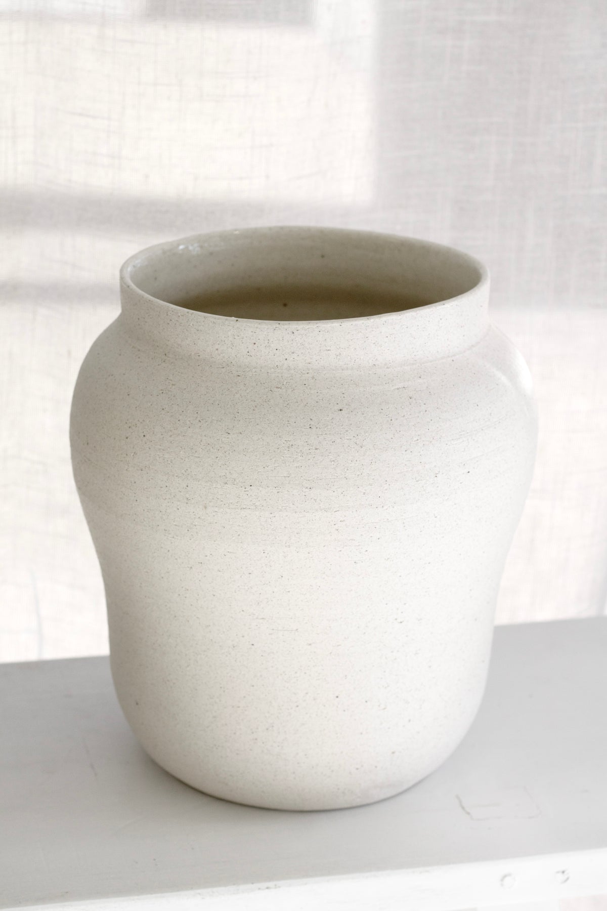 Sandface bellied vase on a white ledge in soft window light, sheer curtain behind
