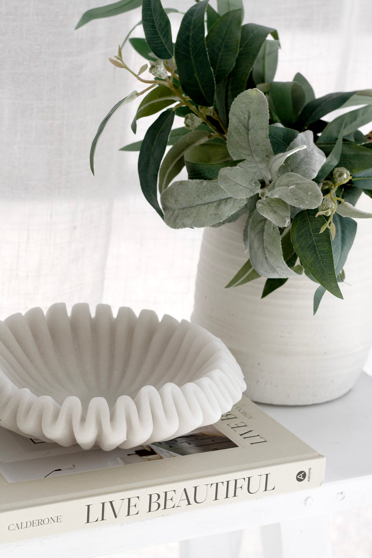 Alma chalk vase with leafy stems and a ruffled bowl on a stack of books by a sheer curtain